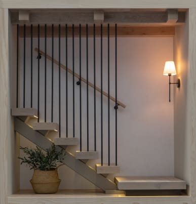 Light oak floating treads on a mono stringer with LEDs, vertical black floor-to-ceiling rods and a wall handrail in a lit nook.