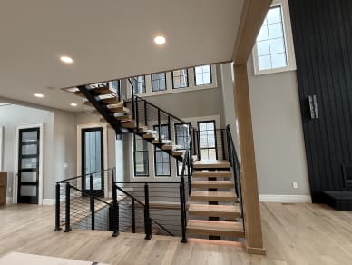 Light oak treads on a black center stringer climb to a landing with black horizontal cable railing beneath tall windows.
