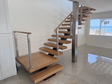 White oak floating treads on a black mono stringer start from a wide bottom landing with stainless cable railing in a bright beach home with marble floors.
