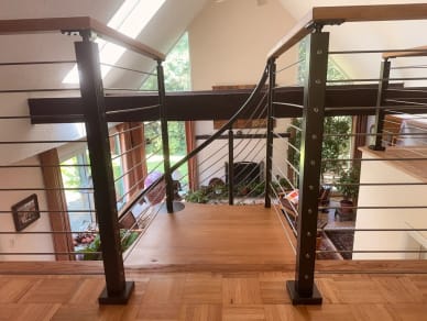Black posts with wood cap and stainless cables guard a parquet loft beside a curving spiral-stair rail in a vaulted room.