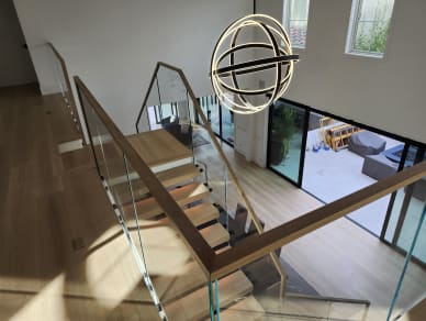 Overhead view of a glass railing with a white oak handrail descending past an orb chandelier to a bright living area.