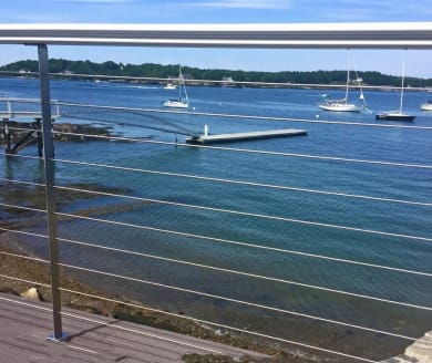 Stainless posts with round handrail and cable infill frame a harbor view with moored sailboats.