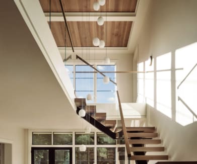 Wood floating treads on a dark mono stringer with frameless glass and a wood handrail climb a sunlit two-story stairwell.