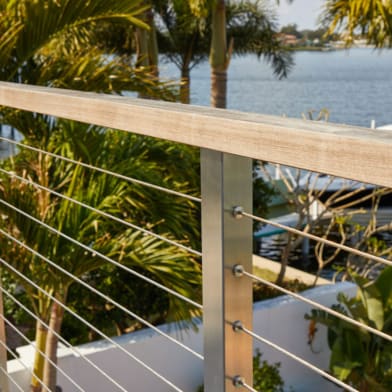 Close detail of a stainless cable post and wood cap rail overlooking palms and a waterway.