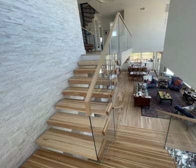 Natural wood floating treads cantilever from a white stacked-stone wall with glass railing and wood handrail above an open great room.