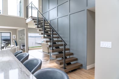 Open wood treads on a black stringer with black post cable railing beside a gray paneled wall in a lake house.