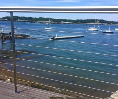 Stainless posts with round handrail and cable infill frame a harbor view with moored sailboats.