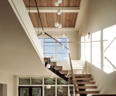 Wood floating treads on a dark mono stringer with frameless glass and a wood handrail climb a sunlit two-story stairwell.