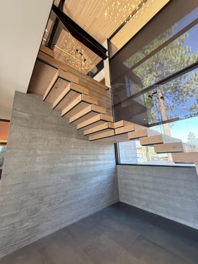 Connected white oak Stack treads float off a board-formed concrete wall beside tall windows framing pines
