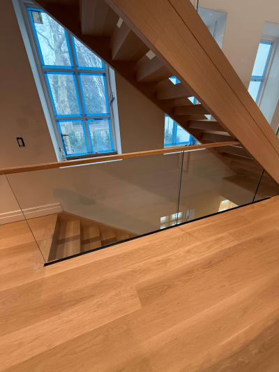 Frameless glass and oak handrail wrap a stair opening below a blue-taped arched window