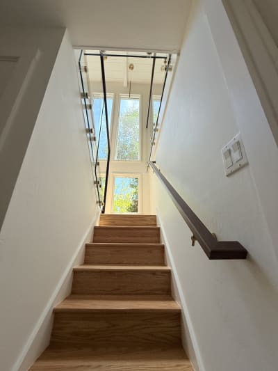 A walnut wall-mounted handrail lines an enclosed oak stair rising toward glass railing and tall windows.