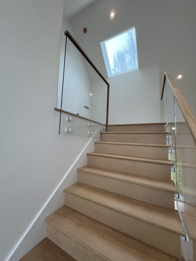 An oak staircase climbs toward a skylight between glass railings on standoff pins with wood handrails.