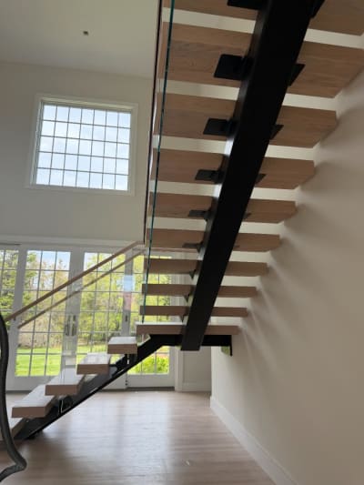 View under a black mono stringer carrying white-oak treads with glass railing in front of tall grid windows.