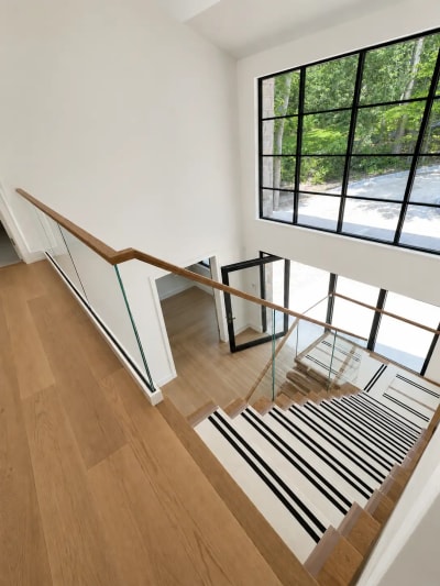 A glass guard with continuous oak handrail wraps a stairwell with covered treads beside large black-grid windows.