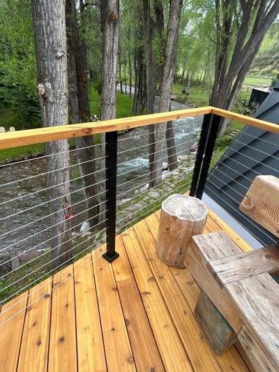 Cedar deck above a rushing creek with black posts, stainless cable infill and a cedar top rail among aspen trees.
