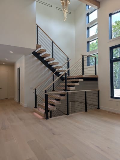 White oak floating treads on a black mono stringer switch back at a landing, with LED under-lighting and black cable railing below tall windows.