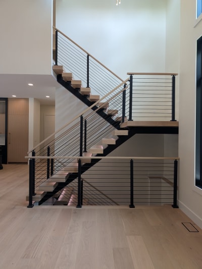 Black mono stringers with white oak treads switch back above a cable-railed basement opening in a bright entry.