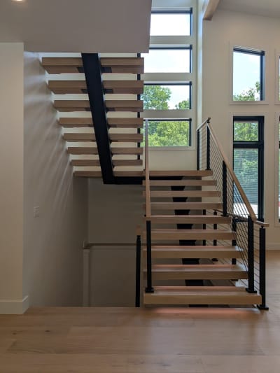 White oak treads on a black mono stringer turn at a landing with cable railing and wood handrail, framed by tall windows.