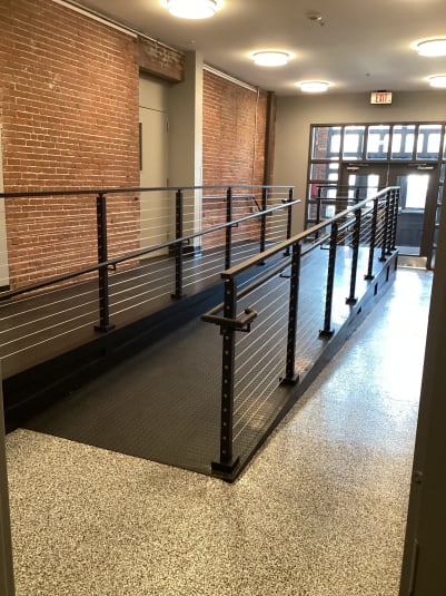 Black posts with cable infill and grab rails line an interior ramp in a renovated brick building.