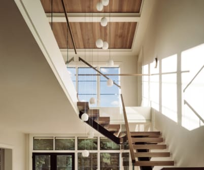 Wood floating treads on a dark mono stringer with frameless glass and a wood handrail climb a sunlit two-story stairwell.