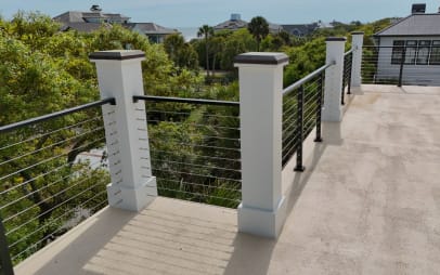 White columns and black cable railing at the corner of an upper deck surrounded by trees.