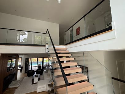 Oak floating treads with a black handrail and frameless glass rising to a glass-railed loft above a living room.