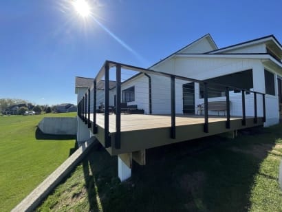 Black slim side-mount cable railing with double corner posts wraps a raised deck on a white farmhouse.