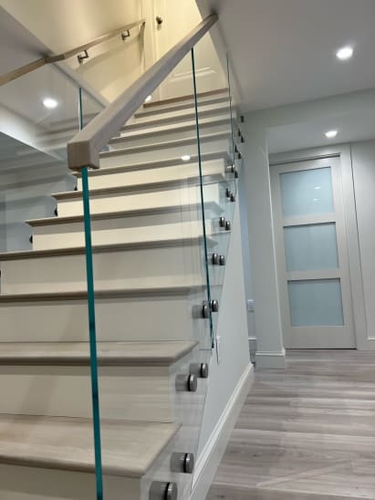 Frameless glass on stainless standoff pins with a light wood handrail along a white closed stair.