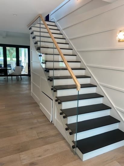 Frameless glass on black standoff pins with an oak handrail beside black treads and white risers.
