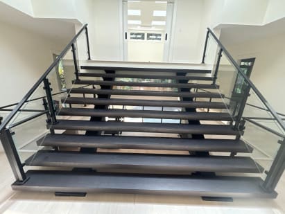Dark wide floating treads on dual stringers with black-post glass railings rise toward a bright skylit hallway.