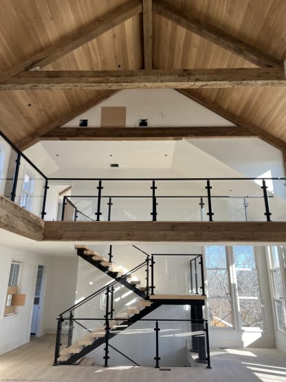 Black mono stringer stair with oak treads and post-to-post glass railing rising to a loft with exposed timber trusses.