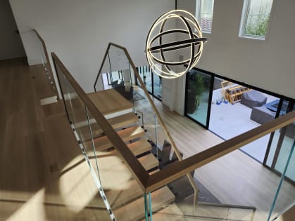 Overhead view of a glass railing with a white oak handrail descending past an orb chandelier to a bright living area.