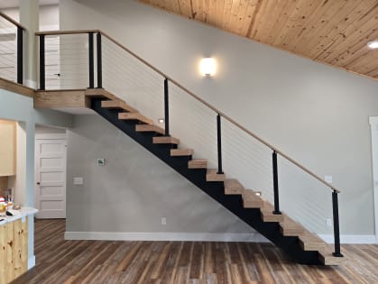 Red oak treads on a black stringer rise to a loft beside black-post cable railing with a wood handrail under a pine ceiling.
