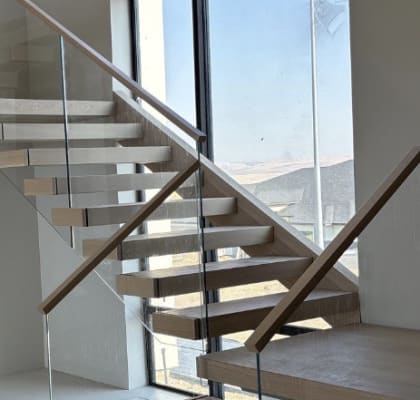 Light oak floating treads turn at a landing with frameless glass and wood handrails beside a tall window.