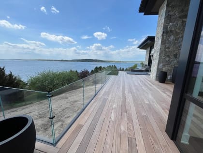 Frameless glass railing runs along a long hardwood deck of a modern stone home with bay views.