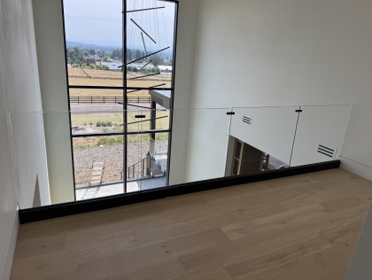 A frameless glass railing set in a black base rail edges a white oak loft overlooking a two-story window.