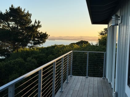 Silver-post cable railing on a wood balcony overlooking trees and water at sunset.