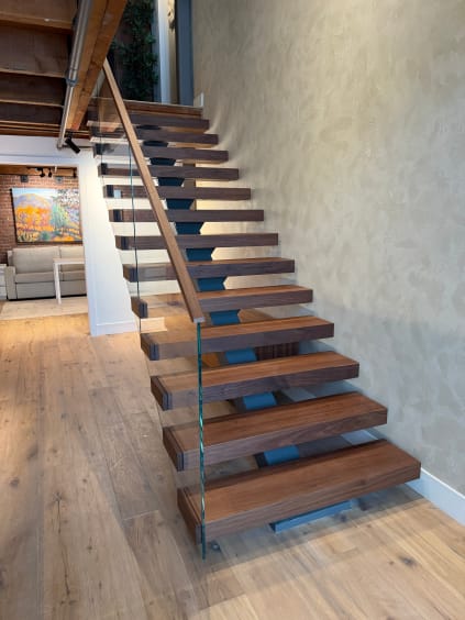 Rich walnut floating treads on a mono stringer with glass railing and wood handrail climb a textured plaster wall.