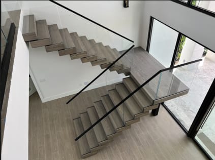 Gray oak cantilevered treads turn at a landing beside frameless glass with a black handrail.