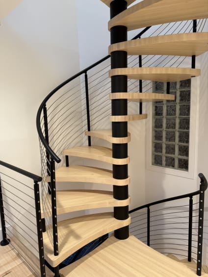 A maple spiral stair winds around a black column, wrapped by a curved black cable railing.