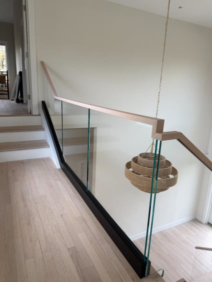 Frameless glass in a black base channel with a squared white-oak handrail guards an open stairwell beside a woven pendant light.