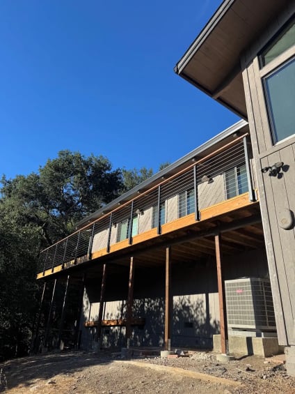 A long elevated wood deck with black posts, wood cap and cable infill runs along a gray home under oak trees.