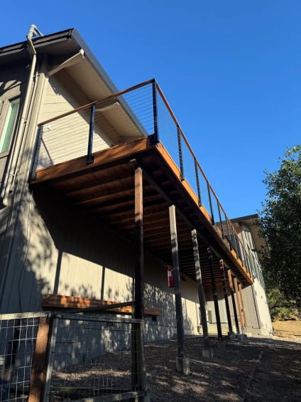 Wood-capped cable railing tops a raised redwood deck on steel posts beside a gray-sided home.