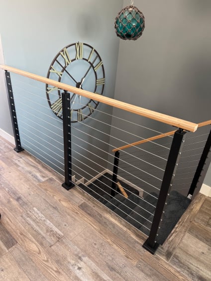Black posts with cable infill and a light wood handrail guard a stair opening on a wood-look floor.