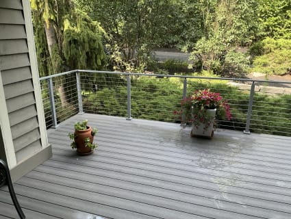 A gray composite deck with flower pots edged by gray cable railing against lush shrubs.
