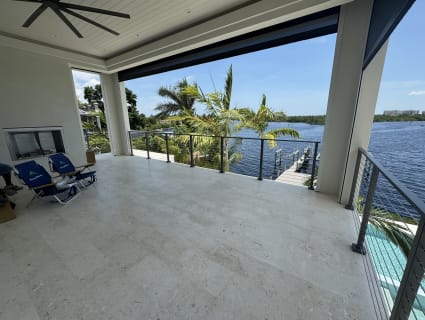 Slim gray rod railing edges a covered stone lanai overlooking a waterway lined with palms.