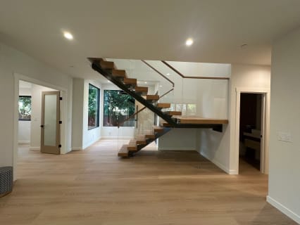 Open-riser floating stair with a black mono stringer, white oak treads and frameless glass railing in a bright modern room.