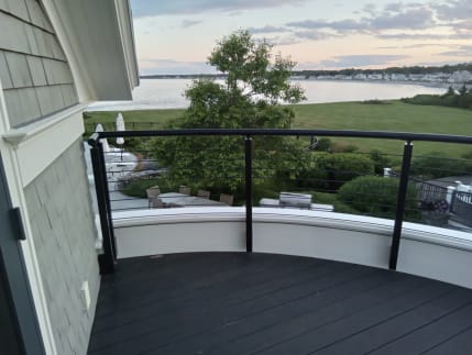 Black curved handrail with cable infill encircles a balcony overlooking a garden and bay.