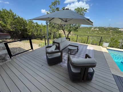 Composite pool deck with lounge seating and umbrella edged by black cable railing above a hill-country view.