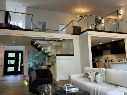 Floating stair with frameless glass leads to a loft lined with glass and wood cap rail above a modern living room; a dog sits on the sofa.